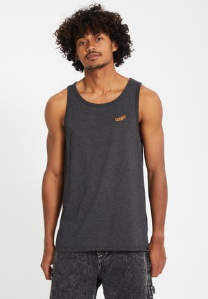 Joven con cabello rizado que lleva una camiseta sin mangas gris oscuro y pantalones cortos de mezclilla lavados con ácido en gris, de pie frente a un fondo liso.