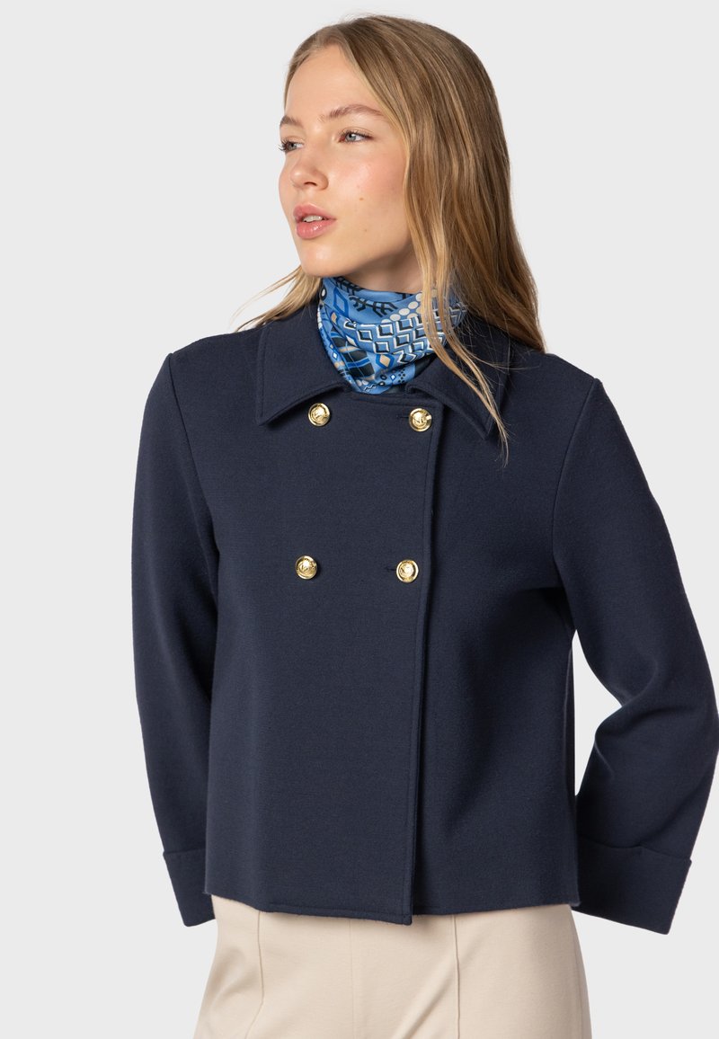 Navy double-breasted jacket with gold buttons, featuring a lapel collar and casual fit. Accompanied by a blue patterned scarf.