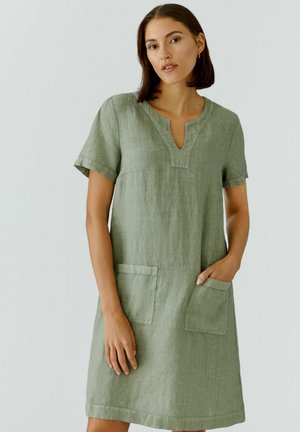 Femme portant une robe en lin vert olive à manches courtes, longueur genou, avec deux poches avant et un décolleté en encoche, debout devant un fond uni.