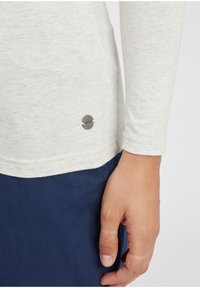 Ljusgrå långärmad skjorta med en mjuk textur och en rund metallisk logotypdetalj på den nedre sidan, i kombination med marinblå byxor.