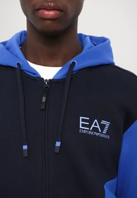 Felpa con zip color navy, con accenti blu e cordino. Presenta un logo EA7 ben visibile sul petto e una texture a costine lungo i bordi.
