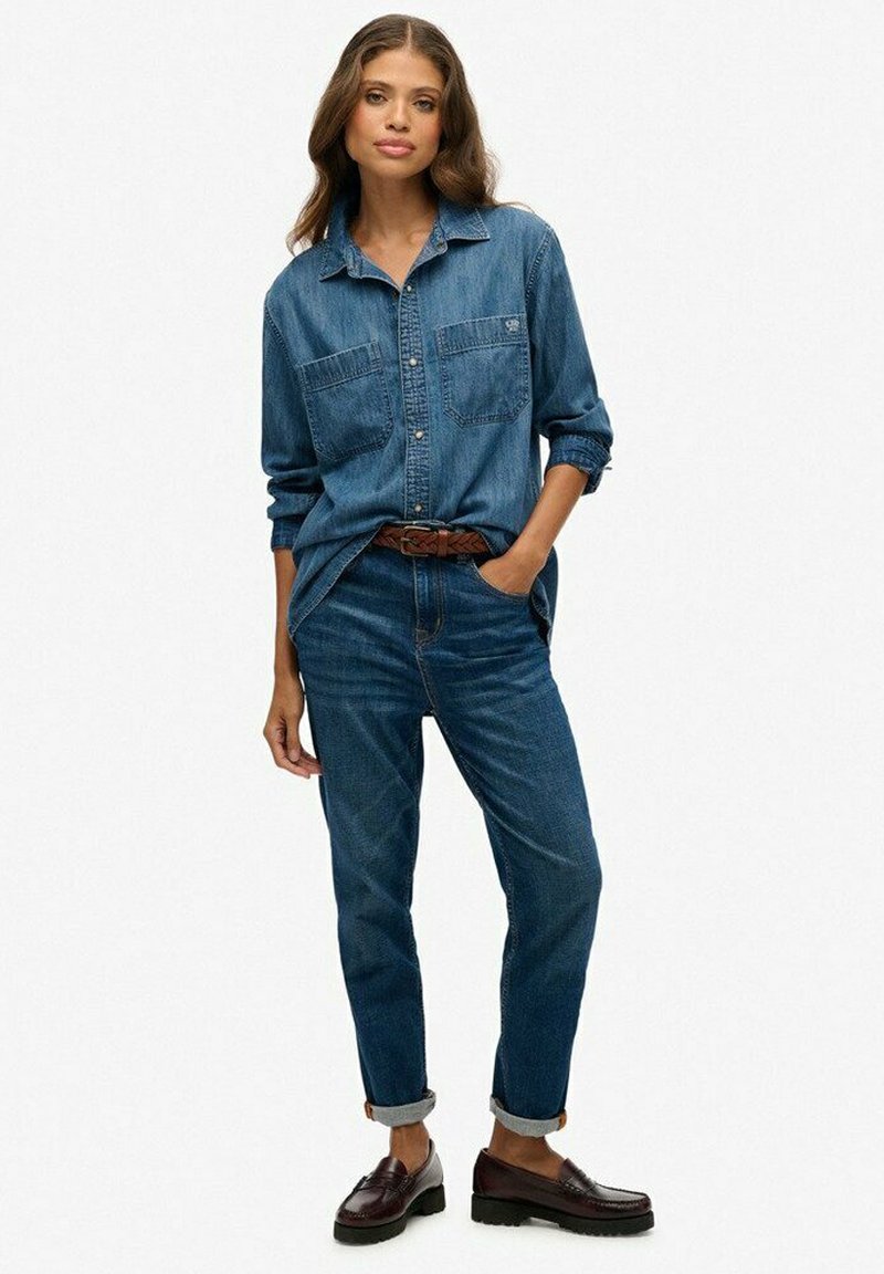 Denim overhemd met lange mouwen en twee borstzakken, gecombineerd met tapered jeans. Donkerblauwe kleur, geweven textuur, bruine riem en zwarte schoenen.