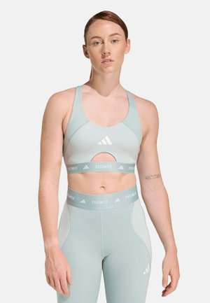 Mujer con sujetador deportivo y leggings Adidas Techfit de color azul claro, de pie contra un fondo blanco liso, mirando hacia adelante.