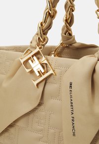 Sac à main beige avec poignées en chaîne dorée, motif embossé, pendentif décoratif avec initiales en or, et foulard en tissu beige imprimé "Elisabetta Franchi."