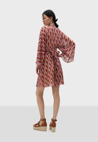 Läpinäkyvä, kuviollinen mekko, jossa on punaisia, sinisiä ja beigejä geometrisiä muotoja. Ominaisuuksiin kuuluvat kellot hihat ja sidottu vyötärö. Yhdistetty ruskeisiin espadrillo-sandaaleihin.