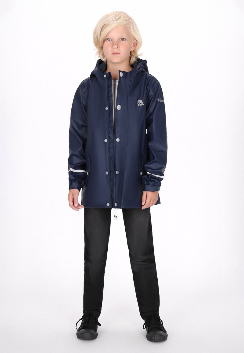 Jūras zila ūdensnecaurlaidīga jaka ar kapuci, spiedpogām, sānu kabatām un pelēkiem akcentiem. Saskaņota ar melniem biksēm un tumšiem sporta apaviem.