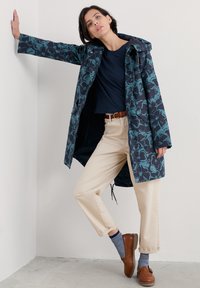 Marineblauwe regenjas met bloemenpatroon, lichtgewicht, met capuchon, in een relaxte pasvorm en met drukknoopsluitingen. Geshakeerd met beige broek en bruine schoenen.