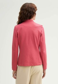 Blazer rose ajusté avec manches longues et col cranté. Tissu texturé avec une finition lisse, présentant des coutures sur mesure et un ourlet net.