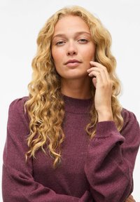 Pull en maille bordeaux avec un col montant, de longues manches bouffantes et une texture douce. Le modèle a des cheveux blonds ondulés et pose de manière réfléchie.