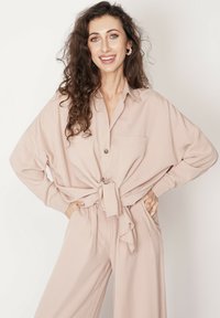 Jasnobeżowa bluzka z wiązanym przodem, w luźnym kroju, z zapiętymi kieszeniami i gładką teksturą, w połączeniu z dopasowanymi luźnymi spodniami.