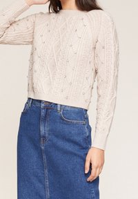 Cremfarbener Kabelstrickpullover mit strukturierten Mustern und dekorativen Perlen. Kombiniert mit einem blauen Jeansrock, der einen entspannten Schnitt zeigt.