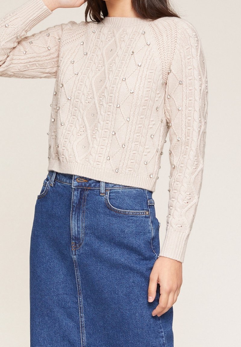 Cremfarbener Kabelstrickpullover mit strukturierten Mustern und dekorativen Perlen. Kombiniert mit einem blauen Jeansrock, der einen entspannten Schnitt zeigt.