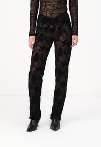 Zwarte, doorschijnende bloemenkant broeken met een relaxed fit, voorzien van een trekkoord in de taille en ingewikkelde bloemmotieven. Gecombineerd met zwarte hakken.