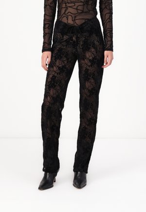 Pantaloni neri in pizzo floreale trasparente con una vestibilità rilassata, dotati di cintura con cordoncino e intricati motivi di fiori. Abbinati a scarpe con tacco nere.