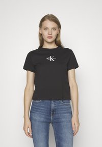 Zwarte katoenen t-shirt met een ronde halslijn en korte mouwen, voorzien van een wit Calvin Klein-logo op de borst. Gepaard met blauwe jeans.
