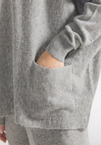 Maglia grigia a maglia con un'ampia vestibilità rilassata, con polsini a coste e una tasca frontale. La texture appare morbida e leggermente pelosa.