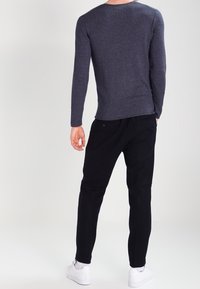 Uomo di spalle che indossa una camicia a maniche lunghe scura, pantaloni neri, calzini bianchi e sneakers bianche, in piedi contro uno sfondo neutro.