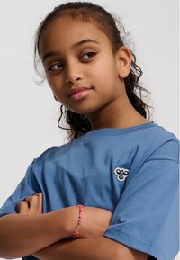 Ung pige med krøllet hår iført blå skjorte krydser armene og ser stille til siden mod en ensfarvet baggrund.