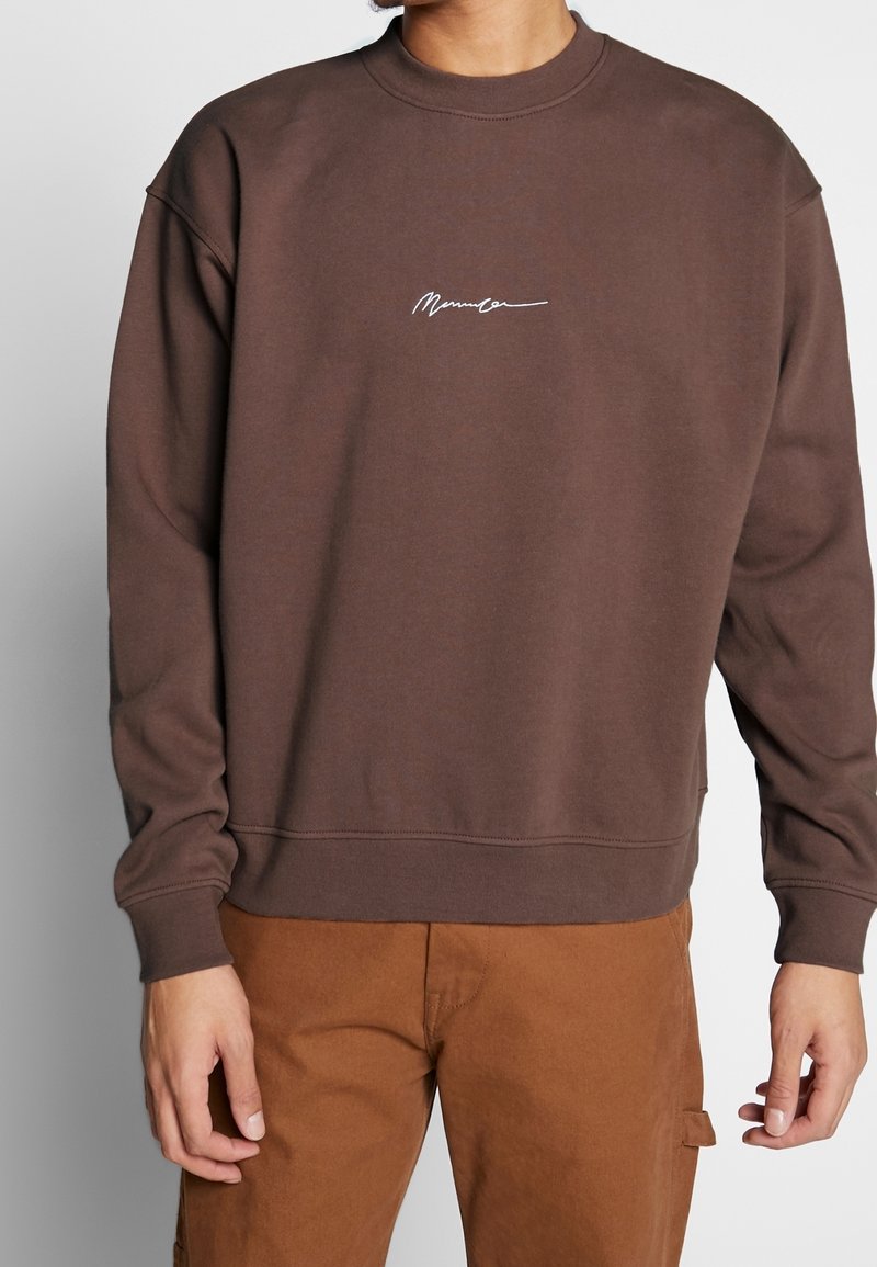 Homme portant un sweat-shirt marron avec un petit texte manuscrit blanc sur la poitrine et un pantalon marron, debout devant un fond uni.