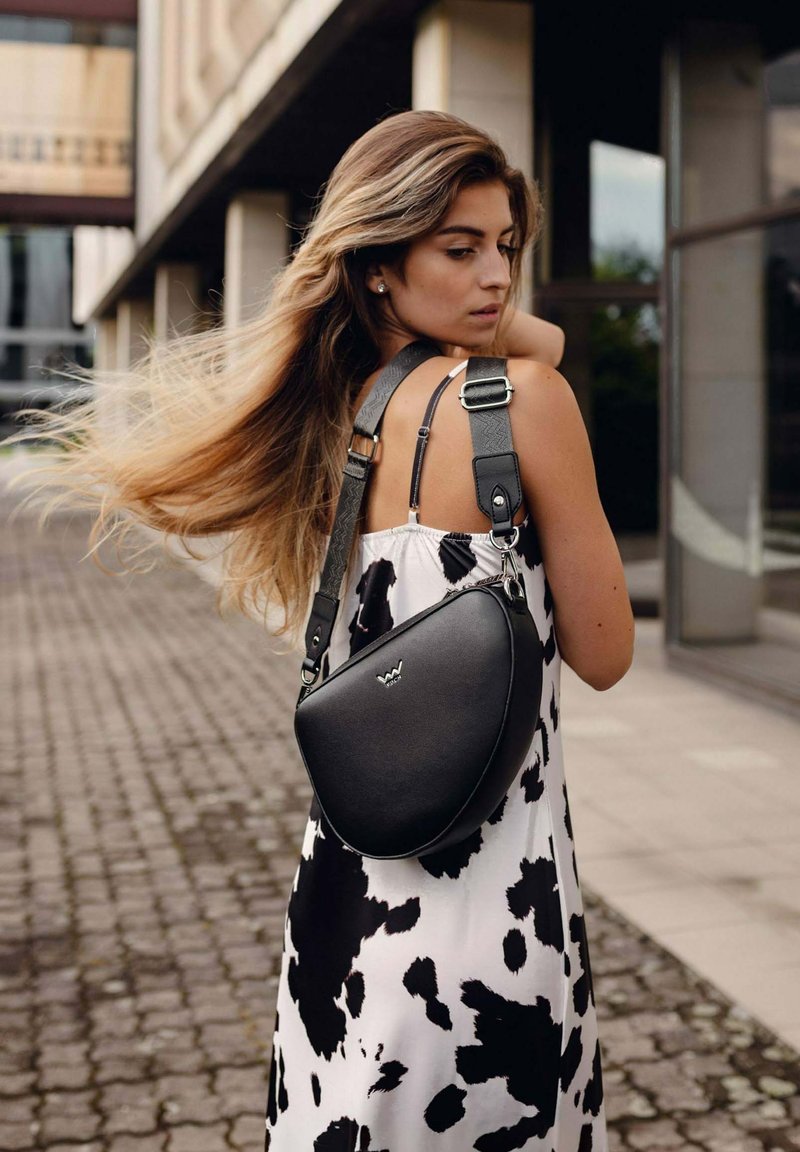 Černá kožená crossbody taška s zakřiveným tvarem, nastavitelným popruhem a kovovými akcenty, nošená přes bílé šaty s černými tečkami.