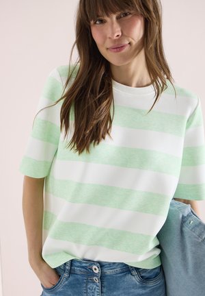 Žena oblečená v krátkém tričku s pruhy mátově zelené a bílé barvy a džíny, držící po straně složený modrý oděv.