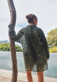 Žena s drdolom, oblečená v priehľadných tmavozelených šatách so zvieracím vzorom, stojaca pri drevenom stĺpe vedľa bazéna, s výhľadom na stromy a hory v pozadí.