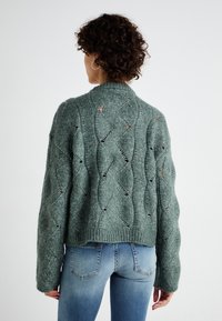 Pullover verde lavorato a maglia con un motivo ondulato e dettagli a giorno, caratterizzato da una finitura a coste e maniche lunghe, indossato sopra jeans azzurri chiari.