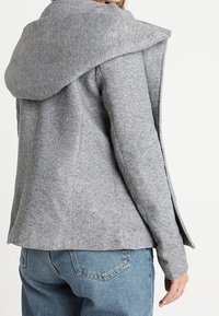 Grå huva jacka med en texturerad finish, som har en avslappnad passform, sidoflikar och en mjuk, lager-på-lager huva design. Kombinerad med blå jeans.