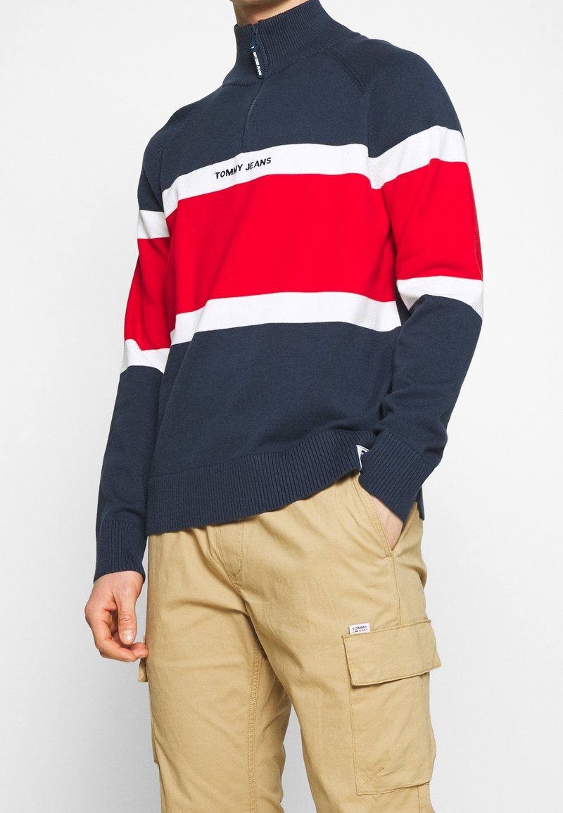 Homme portant un pull Tommy Jeans à rayures bleu marine, rouge et blanc, ainsi qu'un pantalon cargo beige, la main dans la poche, sur un fond uni.