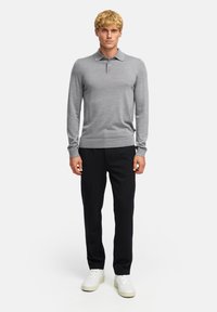 Helle graue Langarm-Polohemd mit Kragen und geripptem Saum. Kombiniert mit schwarzen Hosen und weißen Sneakers, vor einem neutralen Hintergrund.