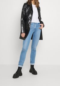 Abrigo negro brillante y largo sobre una camiseta blanca, combinado con pantalones ajustados de mezclilla azul claro y botas de combate negras con cordones y suelas gruesas.