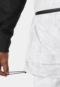 Schwarze Langarmjacke mit einem weiß gesprenkelten Design. Verfügt über Reißverschlusstaschen und einen verstellbaren Kordelzug am Saum.