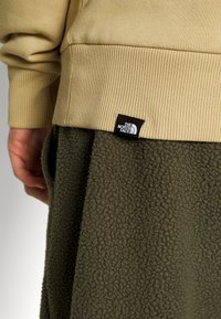 Sweatshirt beige con polsini a coste, abbinato a pantaloni in pile verde scuro caratterizzati da una superficie testurizzata. Il logo The North Face è visibile sul sweatshirt.