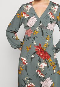 Mujer vestida con un vestido gris con un patrón floral colorido, de mangas largas y escote en V, con las manos en las caderas.