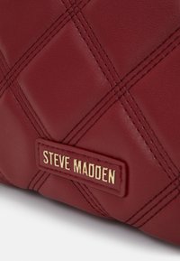 Sac à main en cuir matelassé rouge avec des coutures diagonales et un logo doré "Steve Madden" sur un patch rectangulaire. Texture douce.