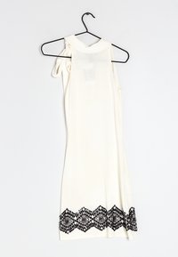 Robe blanche sans manches sur un cintre avec un ourlet en dentelle noire festonnée, présentant un détail de nœud sur une épaule.