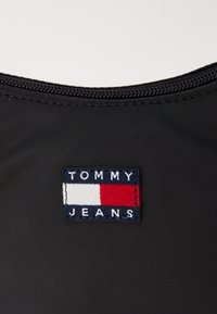 Mustat kankaat, joissa on suorakulmainen brodeerattu kohdemaalaus "TOMMY JEANS"; vaaleansininen, punainen ja valkoinen värilohko. Vetoketjuyksityiskohta näkyvissä.