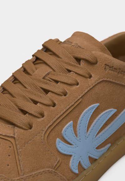 Καφέ σουέντ αθλητικό παπούτσι με χοντρές κορδόνια, ένα ραμμένο μπλε λογότυπο σε σχήμα φύλλου στο πλάι και το όνομα της μάρκας ανάγλυφο κοντά στον γιακά.