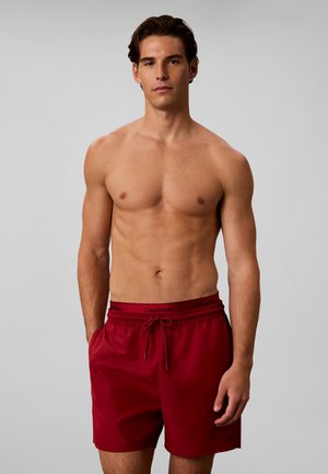 Hombre sin camiseta con cabello oscuro que lleva pantalones cortos de baño rojos Calvin Klein, de pie contra un fondo gris liso.