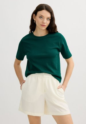 Mujer con cabello castaño ondulado que lleva pendientes de aro dorados, una camiseta verde oscuro de manga corta y pantalones cortos blancos y sueltos con las manos en los bolsillos.