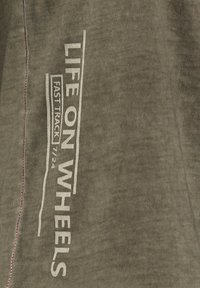 Tela verde oliva con una textura suave; presenta texto impreso en blanco "LIFE ON WHEELS" y "FAST TRACK 7/24" a lo largo del lado, acentuado por costuras rojas.