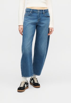 Oseba oblečena v modre široke kavbojke, bele nogavice, črno-bele superge in krem pulover na prsnem delu, stoji pred enostavno belo ozadje.