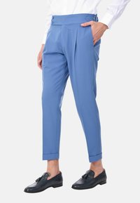 Pantaloni tapered celeste con pieghe frontali, vita con due bottoni, tasche laterali e risvolti, indossati con mocassini neri.