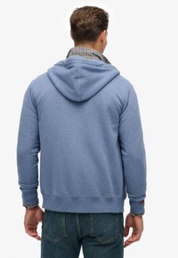 Haut à capuche bleu en tissu doux avec un cordon de serrage, des poignets côtelés et un ourlet. Présente un col à carreaux qui dépasse de dessous.