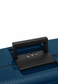 Valigia rigida blu con texture opaca, manico nero e pannello di controllo con porte audio e di reset. Include una serratura di sicurezza notevole.
