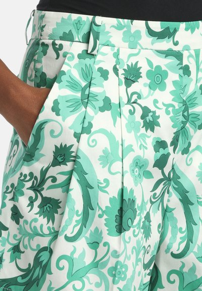 Main dans la poche d'une jupe blanche à imprimé floral vert et bleu sarcelle, avec des plis et une ceinture à passants.