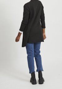Cardigan lungo nero con texture, dalla vestibilità comoda e spacchi laterali, abbinato a jeans blu a lunghezza caviglia e stivali neri con tacco spesso.