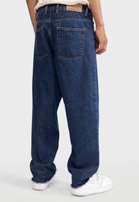 Jean en denim bleu avec une coupe ample, dotés d'une jambe droite, de deux poches arrière et de détails d'ourlet effiloché. Associés à des baskets blanches.