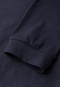 Laivansininen pitkähihainen vaate, jossa on sileä ja hienojakoinen kangas. Hihansuissa on tiivis resori ja hienovaraisia ompeluyksityiskohtia.