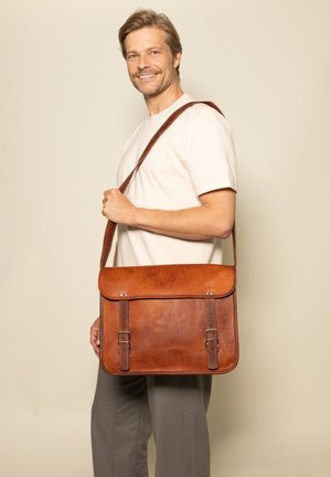Brown leather messenger bag with flat design, adjustable strap, two buckled closures, and smooth texture, held by a person in casual attire.